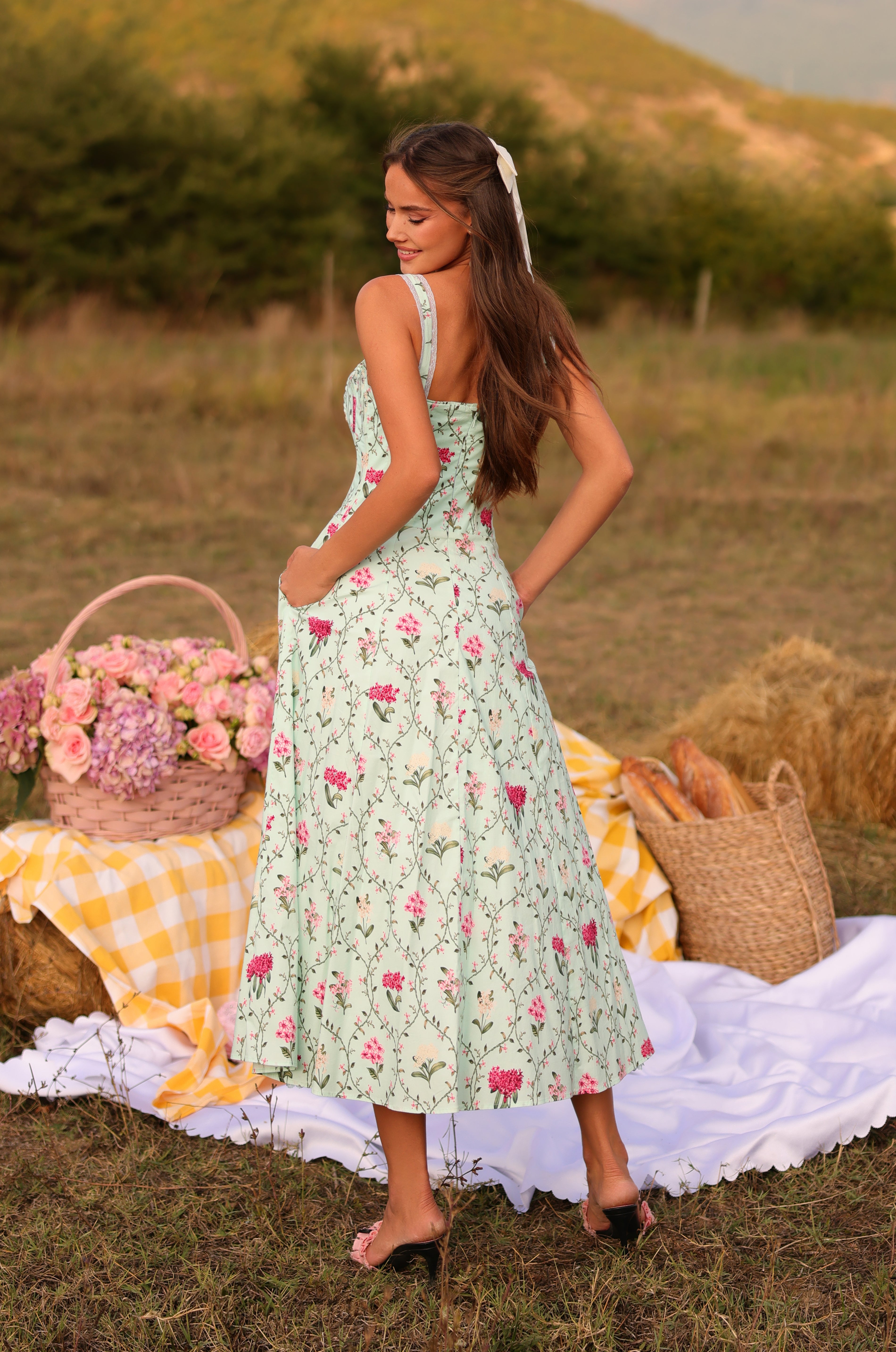 A woman in the Mint Green Floral Casual Midi Dress: Celenia stands on a picnic blanket outdoors, surrounded by a basket of flowers and bread on patterned fabric.